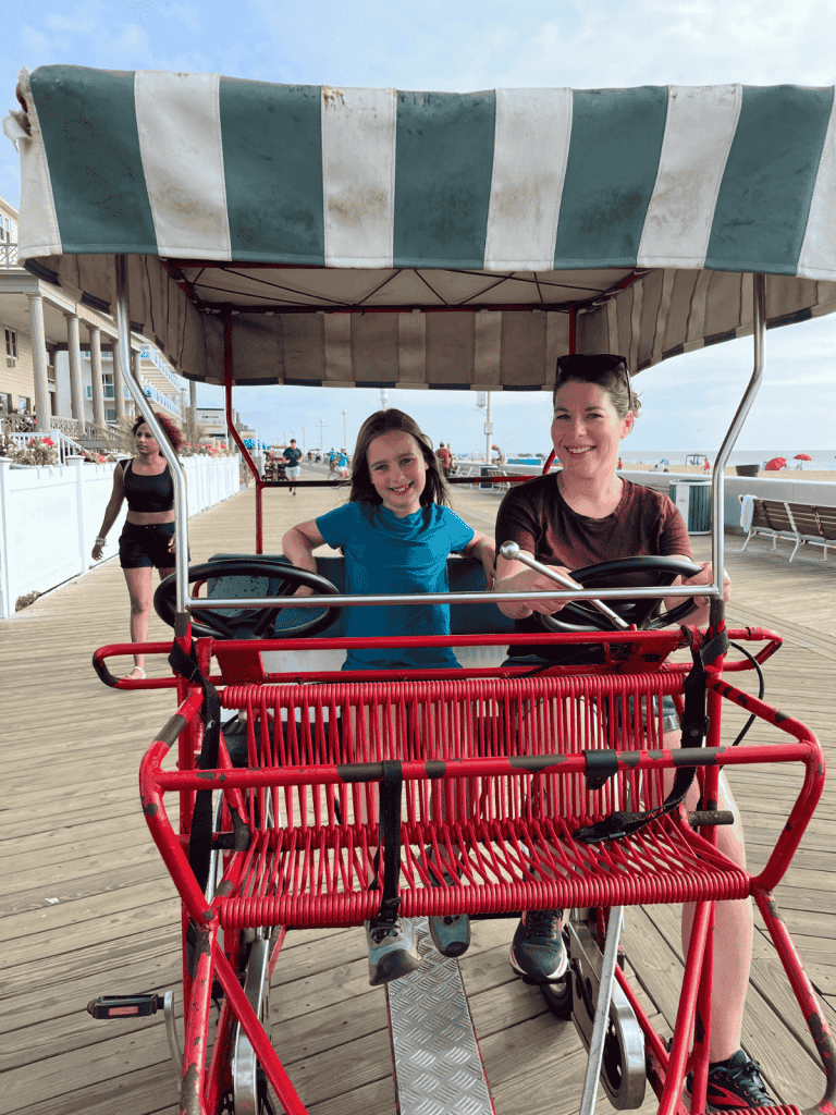 A mom and daughter smile while riding on a surrey bike on the boardwalk with the beach in the background.