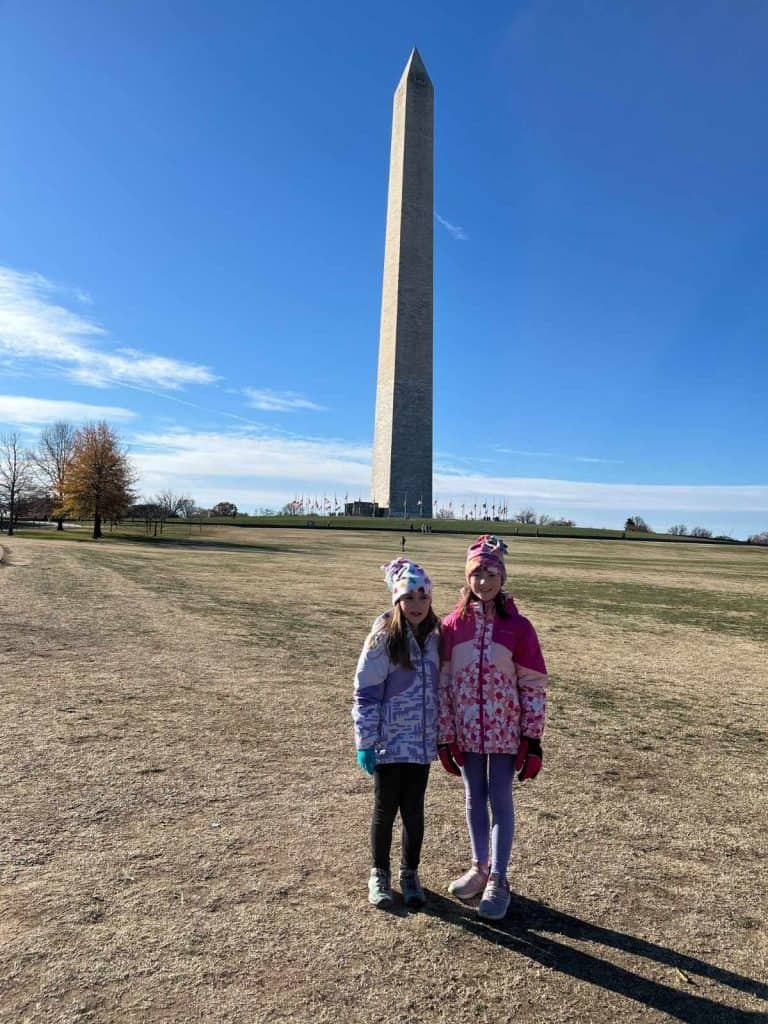 Two children standing at the base of the Washington Monument in D.C. under a clear blue sky.