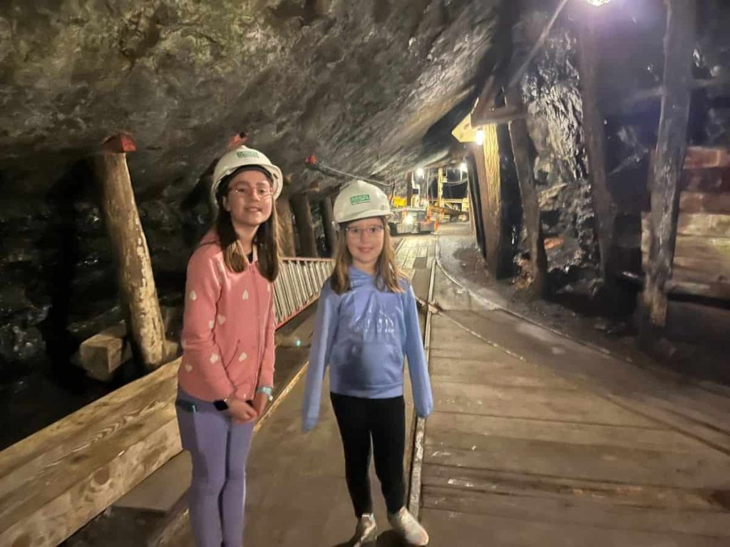 Two girls stand inside a lit coal mine with hard hats on.