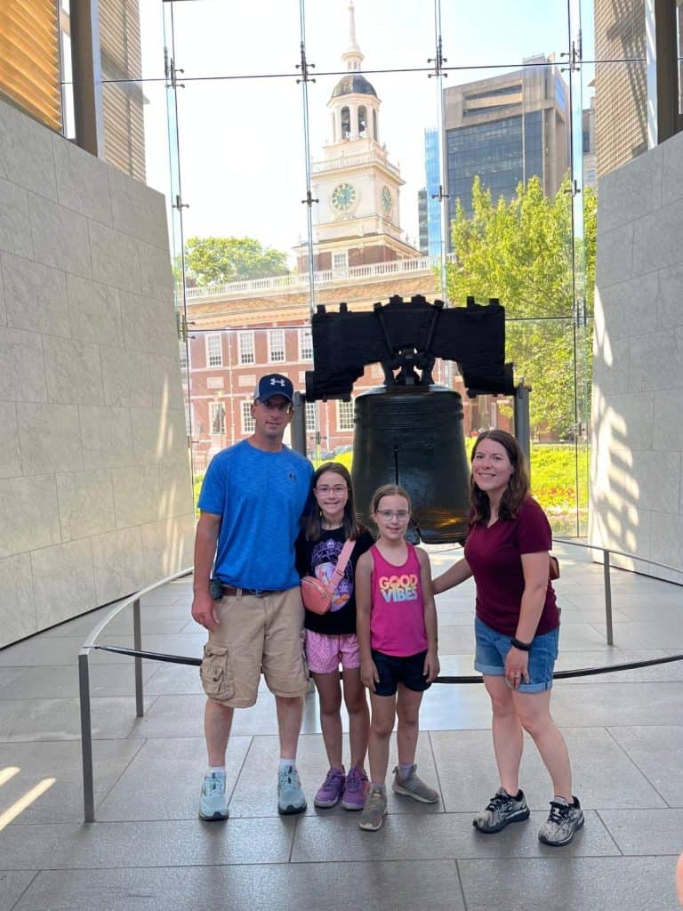 A family of four stands in front of the Liberty Bell with Independence Hall behind them.