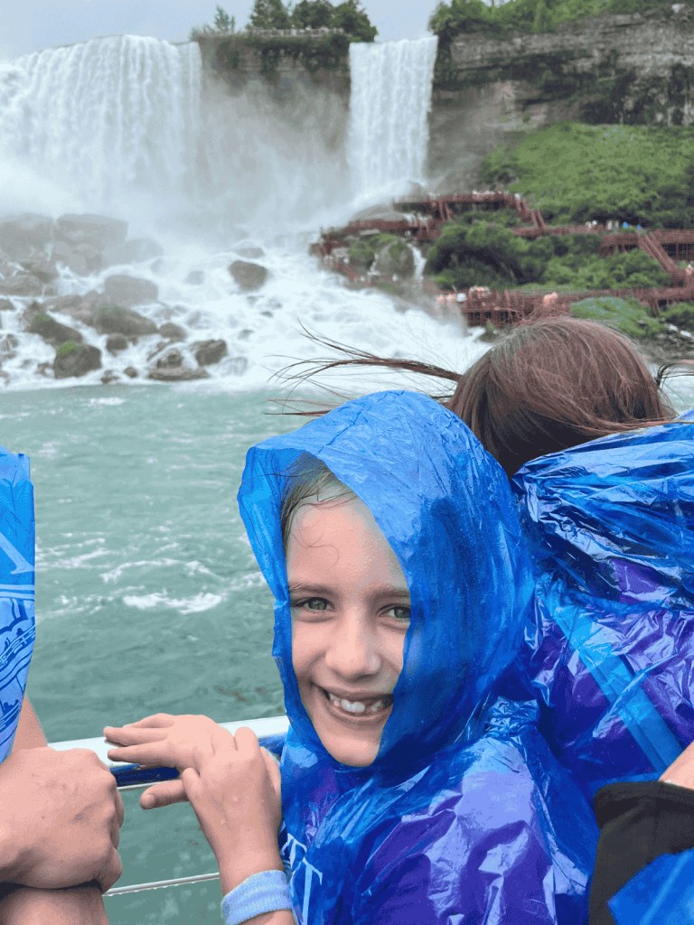 A girl in a blue poncho smiles next to a boat railing with Niagara Falls waterfalls in the background.