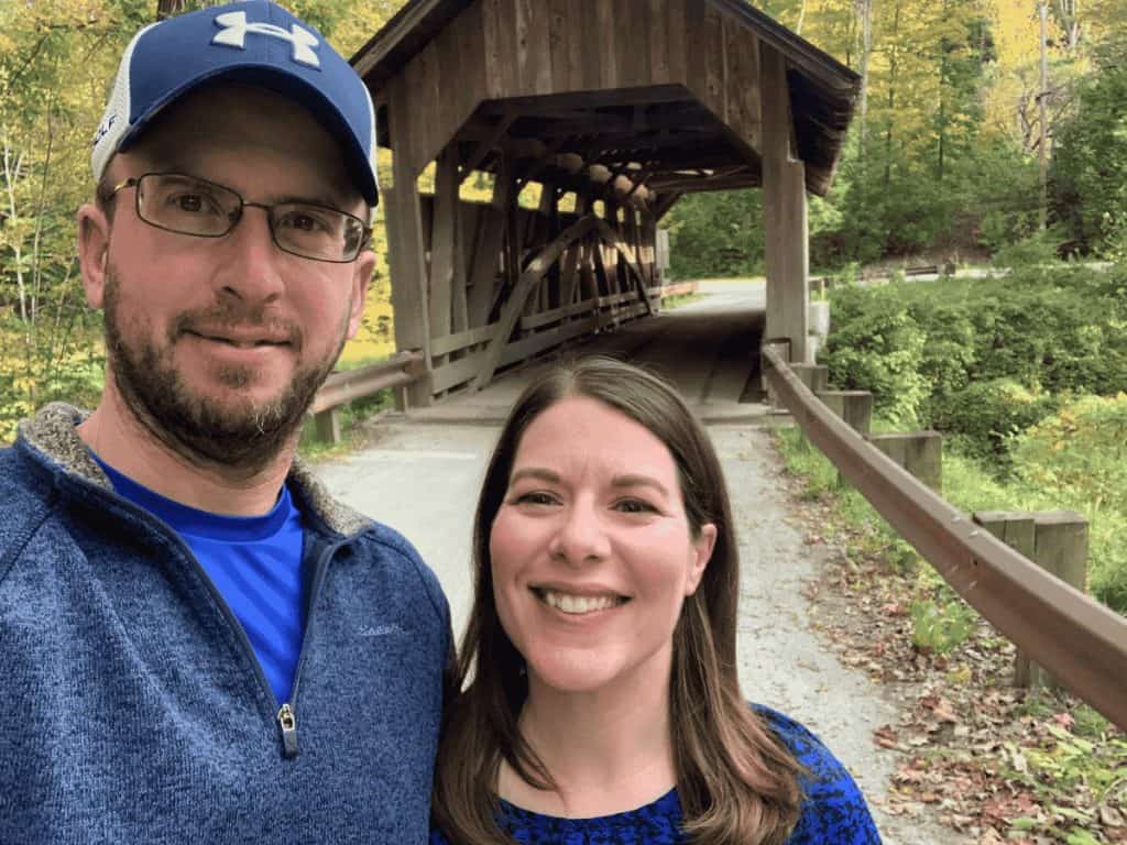 A couple standing in front of a covered bridge and foliage in Vermont.