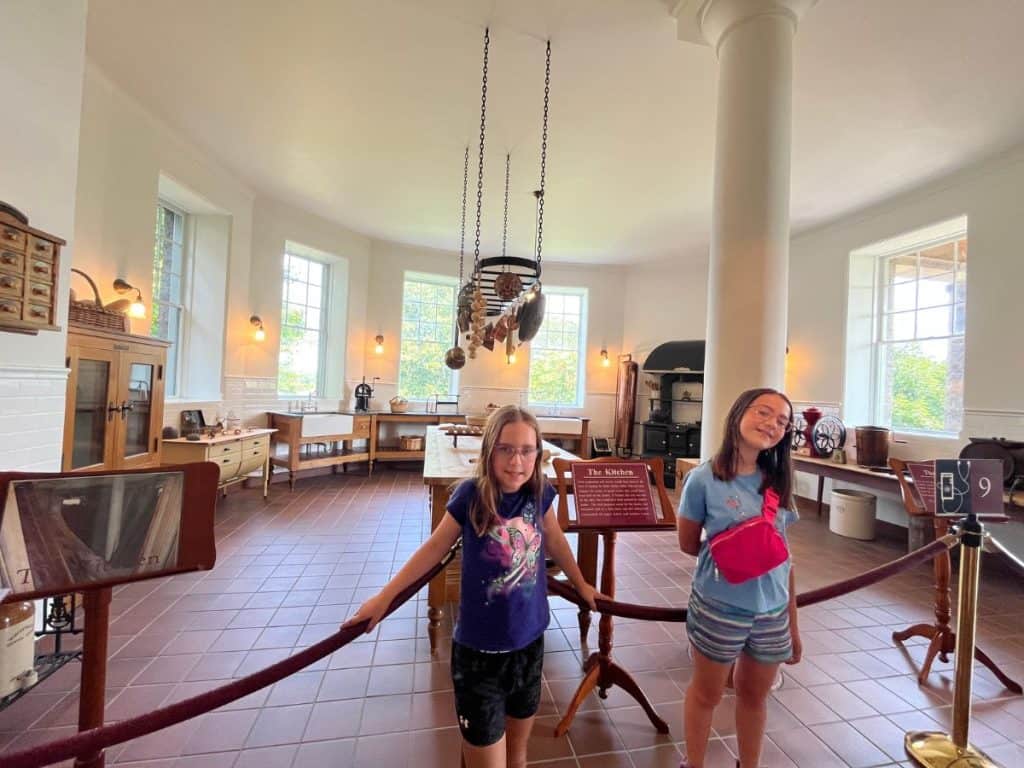 Kid-friendly stop inside Boldt Castle on a 1 day family itinerary Children posing in front of a rope that is in front of a castle's kitchen with old fashioned furniture.