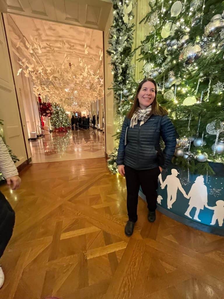 A woman smiling and standing near a decorated tall Christmas tree near a White House hallway on a tour.