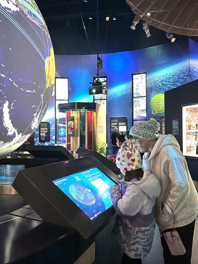 A woman and girl stand at a museum looking down at a touch screen that is in front of a large projection globe.