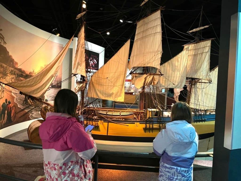 Two girls face a museum display of historical ships with sails in pink and blue coats.