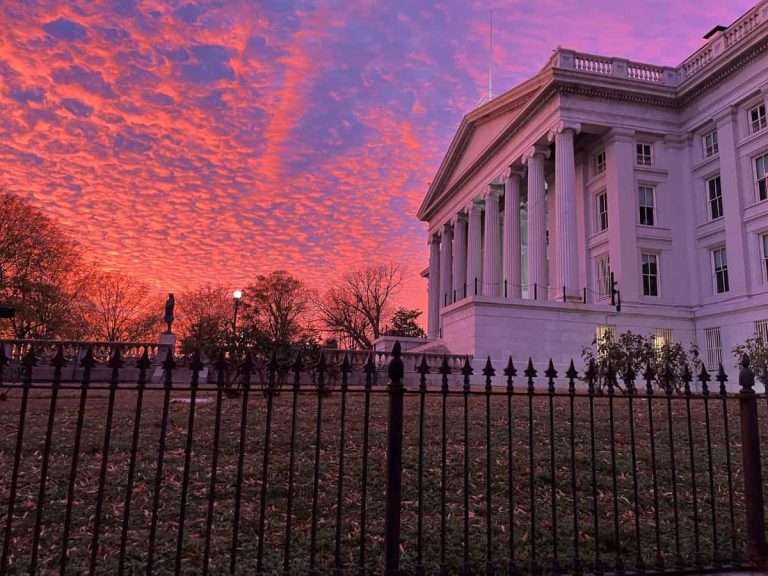 A white government building in Washington DC with a green lawn and iron gate is pictured with a bright colorful sky at dusk.