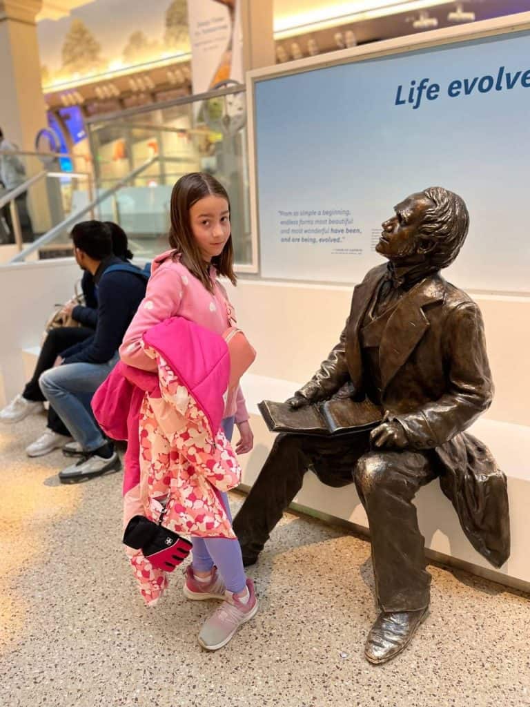 A girl with her eyebrows raised looks at the camera next to a statue in a Smithsonian museum that is sitting looking at her with a book.