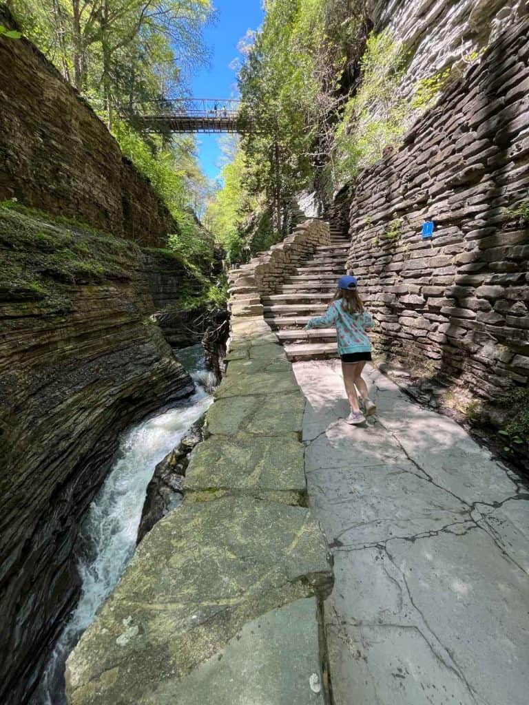 Shows our family on a day trip walking on the path at Watkins Glen State Park. A girl walking on a stone path towards steps and near water rapids on a day trip to Watkins Glen State Park.