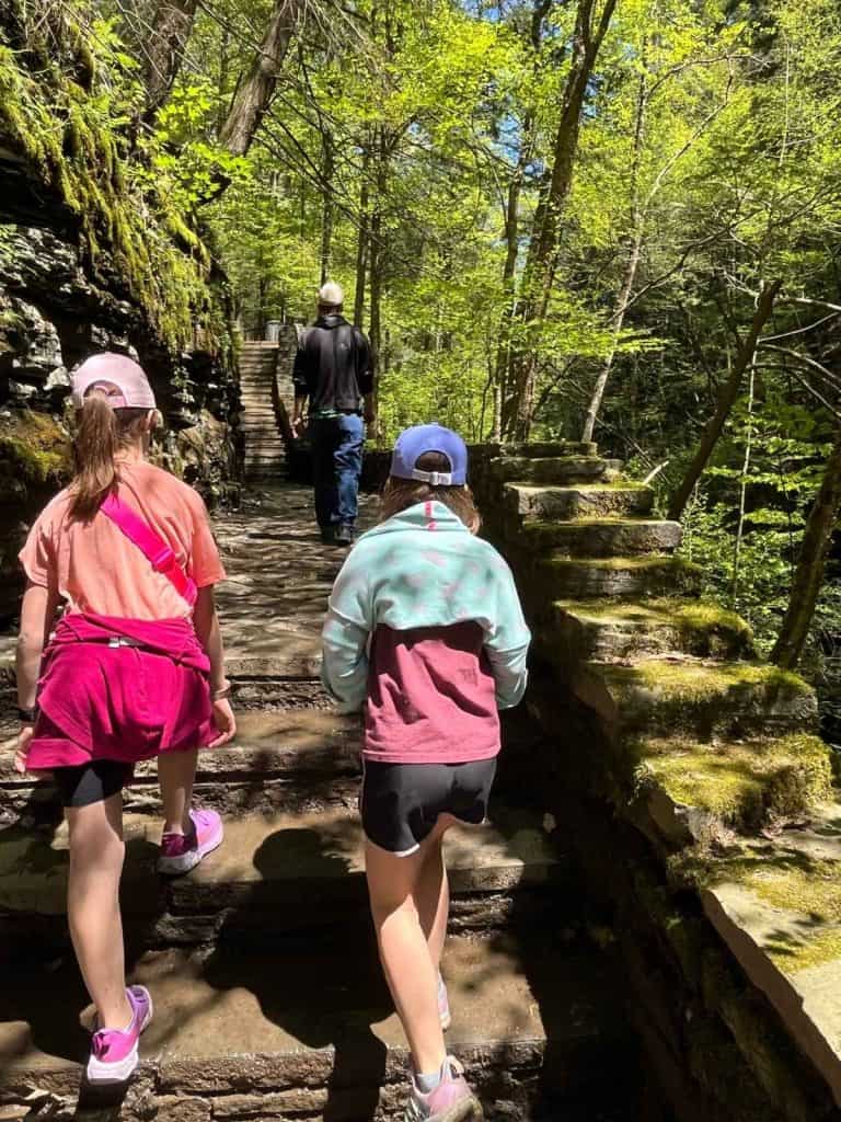 Kids hike on the Watkins Glen path on a family day trip. Two girls in shorts, t-shirts, and hats, hike on a stone stairway and path in a wooded, mossy area.