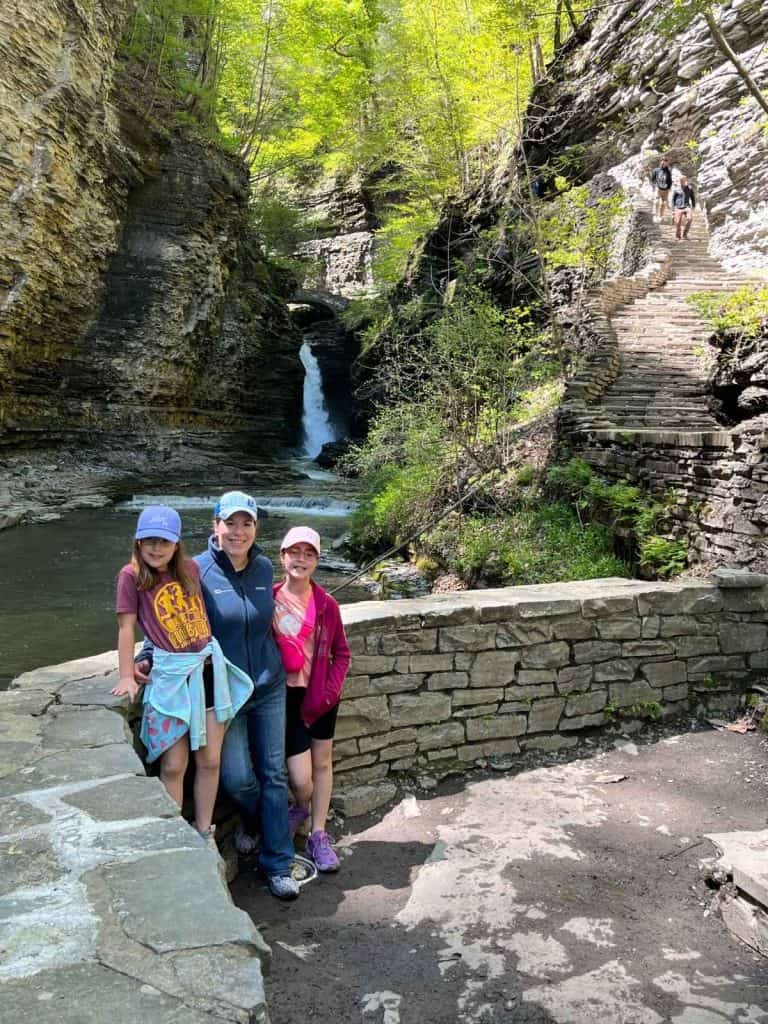 A woman and two kids on a family day trip to Watkins Glen state park for a hike. A woman and two kids stand in front of a circular stone half-wall, with stone stairs, a waterfall, and rapids in the background.