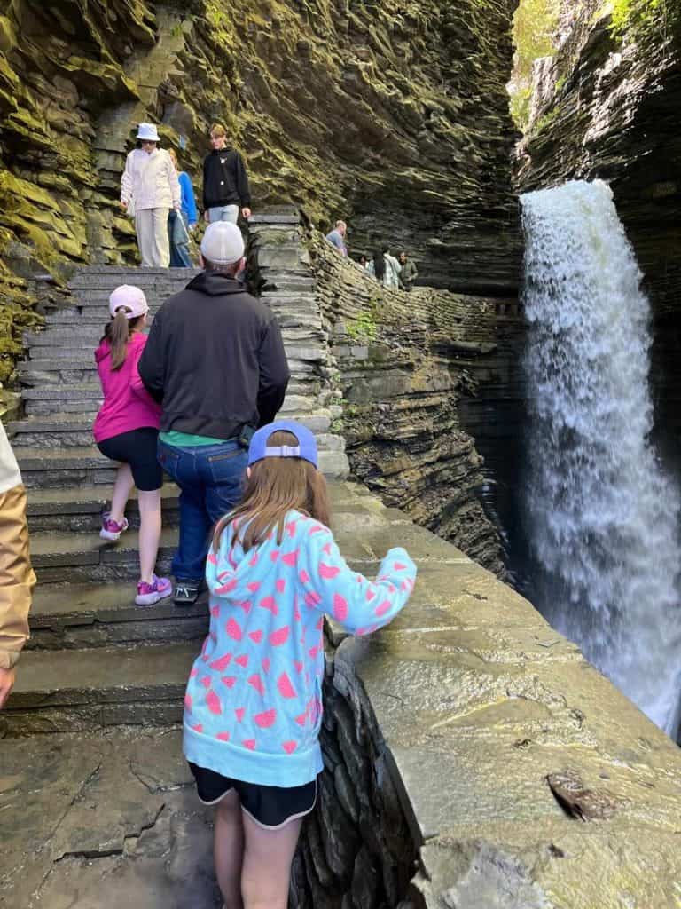 A family hikes up stone steps near a waterfall at Watkins Glen. A dad and two girls walking up stone steps while people are coming down the top landing of stone steps, in front of a rushing waterfall.