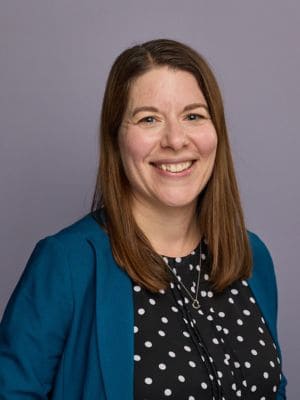 A brunette woman, Amanda Cave Jackson, poses in front of a gray background wearing a black shirt with polka dots and a turquoise jacket.