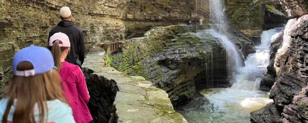 A family of 3 walks toward a cascading waterfall on a stone walkway through a gorge in Watkins Glen, NY.