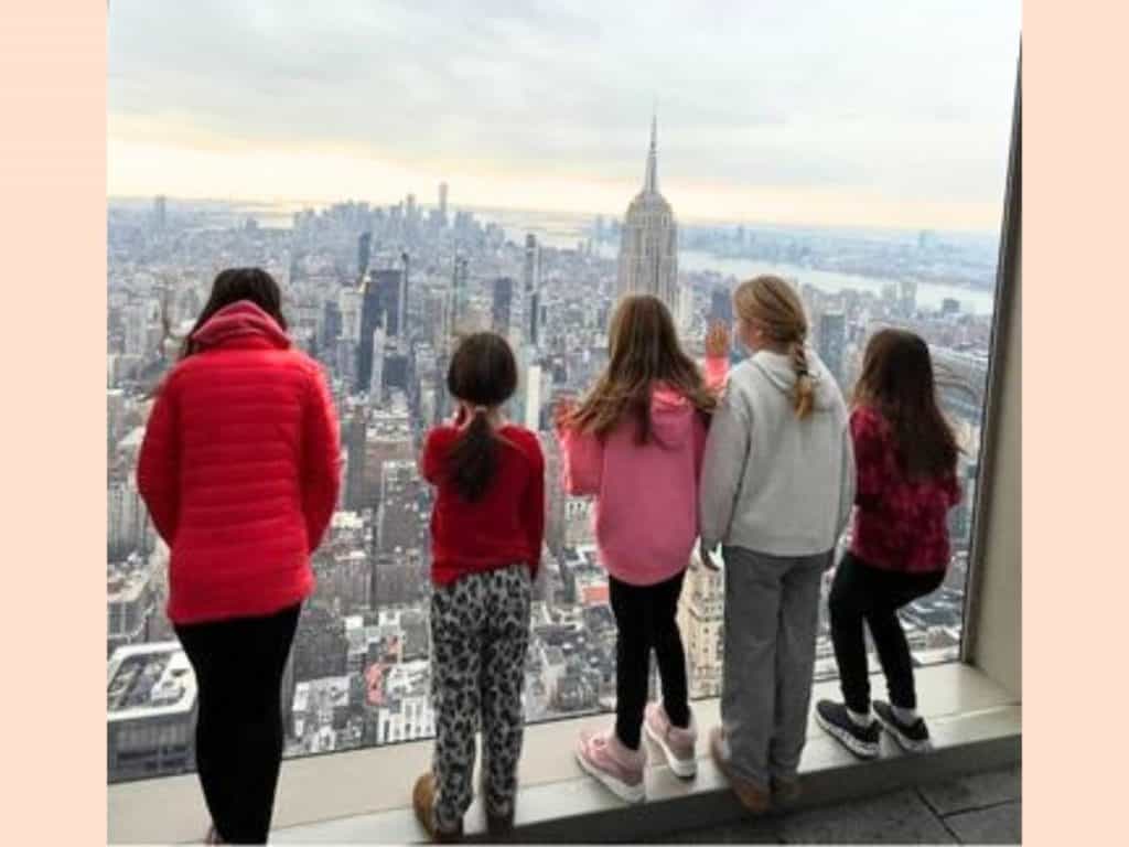 Kids on a New York City guided small group trip at Summit One. Five girls face out at the NYC skyline in wonder through a floor to ceiling window on a cloudy day.