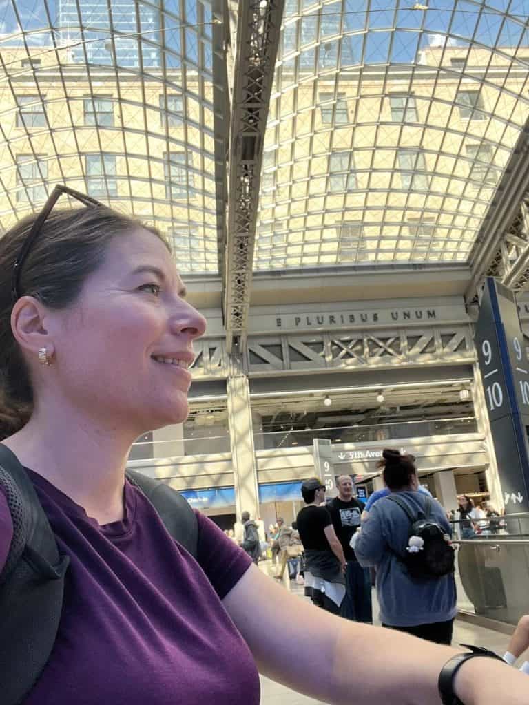 A woman smiles in a selfie at the modern Moynihan Train Hall in NYC, featuring the iconic vaulted glass ceiling and historic 'E Pluribus Unum' inscription in the background.