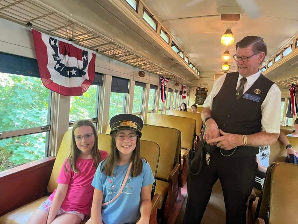Two girls on a restored Cooperstown train next to the conductor, with one wearing his hat and smiling.