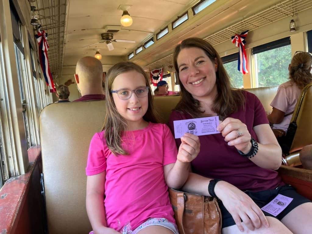 A mom and girl seated on the train, smile and show off a purple souvenir train ticket.