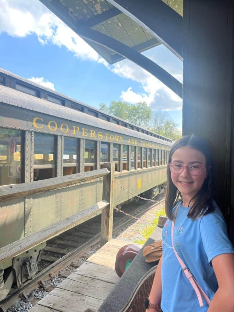 A girl stands smiling on a train platform with the Cooperstown and Charlotte Valley Railroad train in the background.
