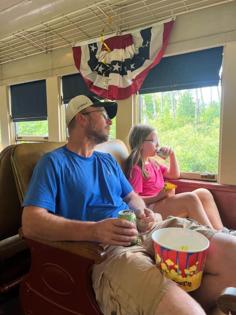 A father and daughter sit in coach train seats with popcorn and snacks in their laps.