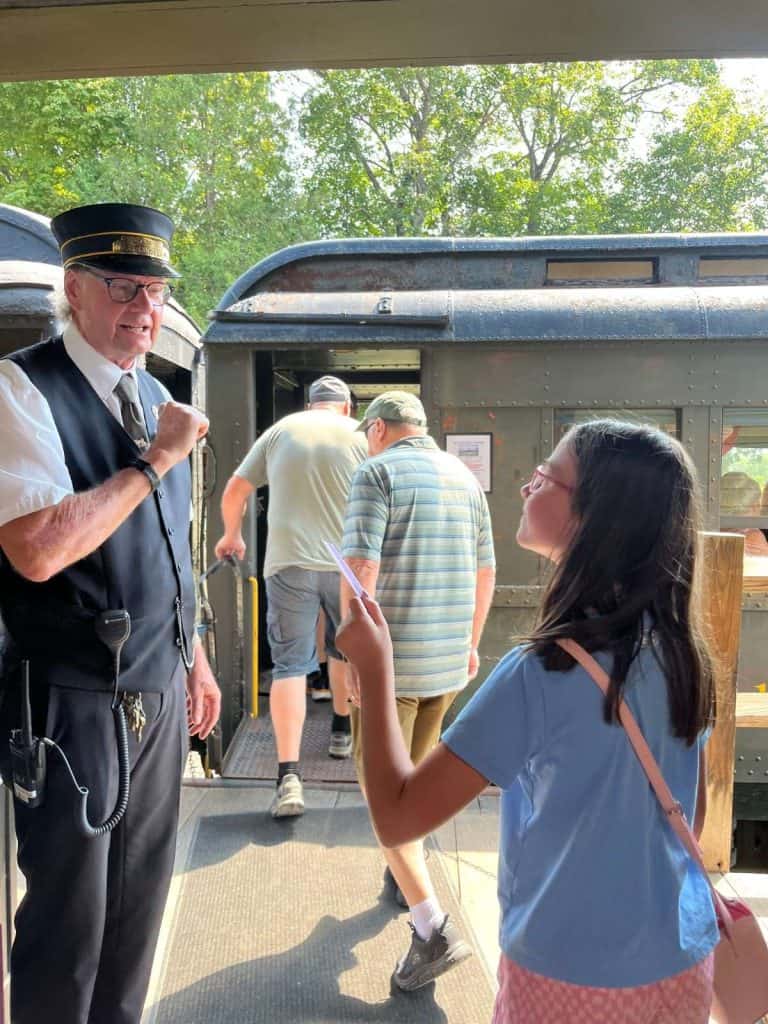 A girl is smiling giving her train ticket to a conductor, with others boarding the train in front of her.
