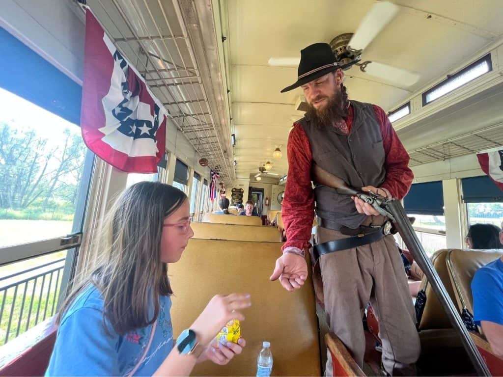 A train robber with gun has their hand out to a girl seated with candy.