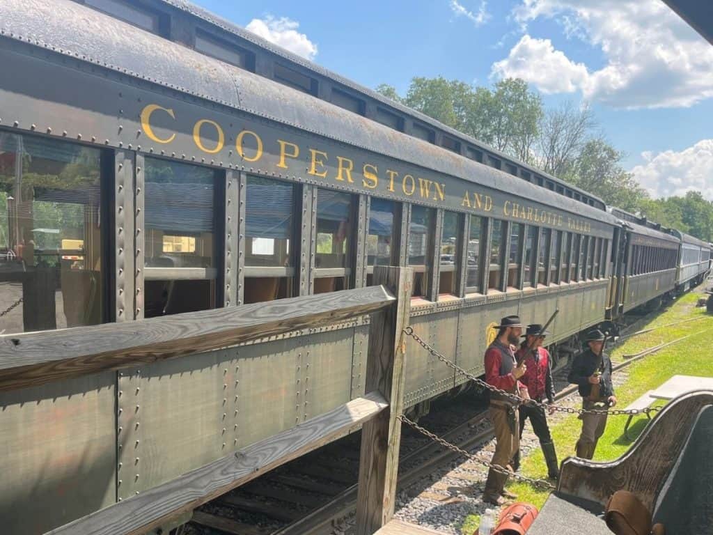 Three train robbers stand in the grass in front of the Cooperstown and Charlotte Valley train with guns ready.