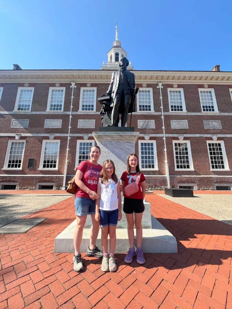 A family standing in front of the brick Independence Hall building in front of a George Washington marble and bronze statue.