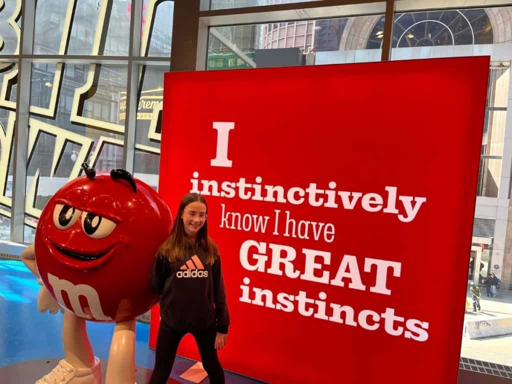 A girl stands beside a life-size red M&M character and next to a bright red poster that says I instinctively know I have great instincts.