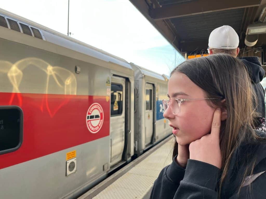 A family stands on a train platform in NYC. A girl plugs her ears on a NYC train platform as the train arrives.