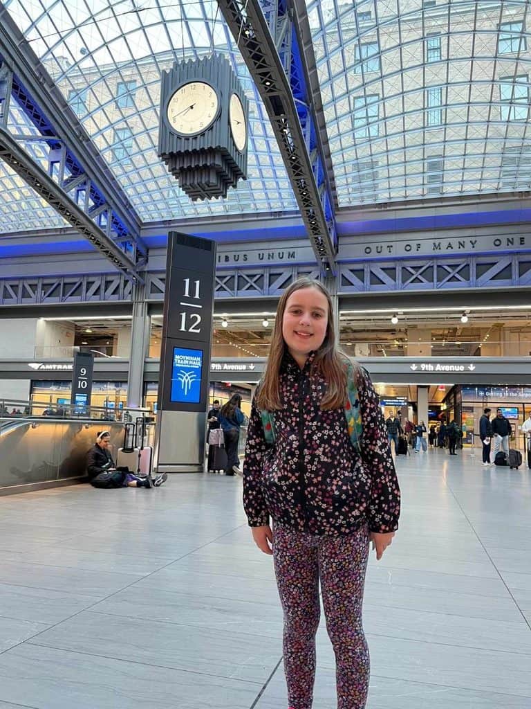 A family at Penn Station on their first trip to NYC. A girl smiles as she stands in Penn Station train station by the 11/12 platform sign and below a clock and vaulted ceiling.