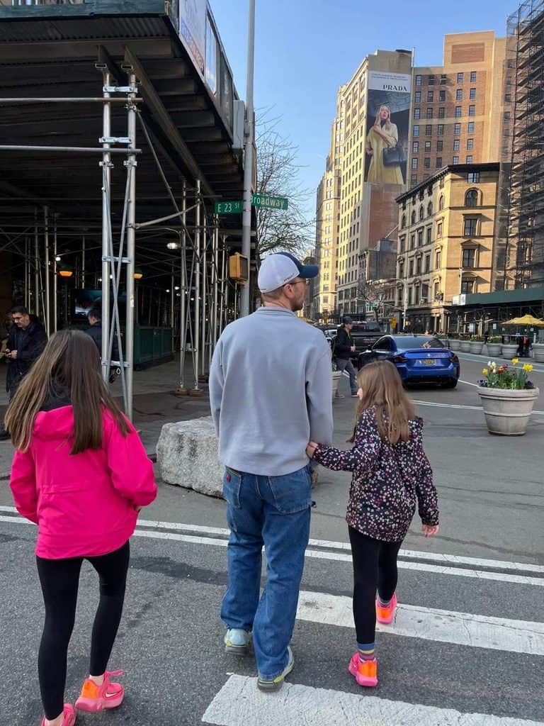 Families will walk a lot on a first trip to NYC. Two girls and their dad walk across a NYC sidewalk with high rise buildings and scaffolding nearby.