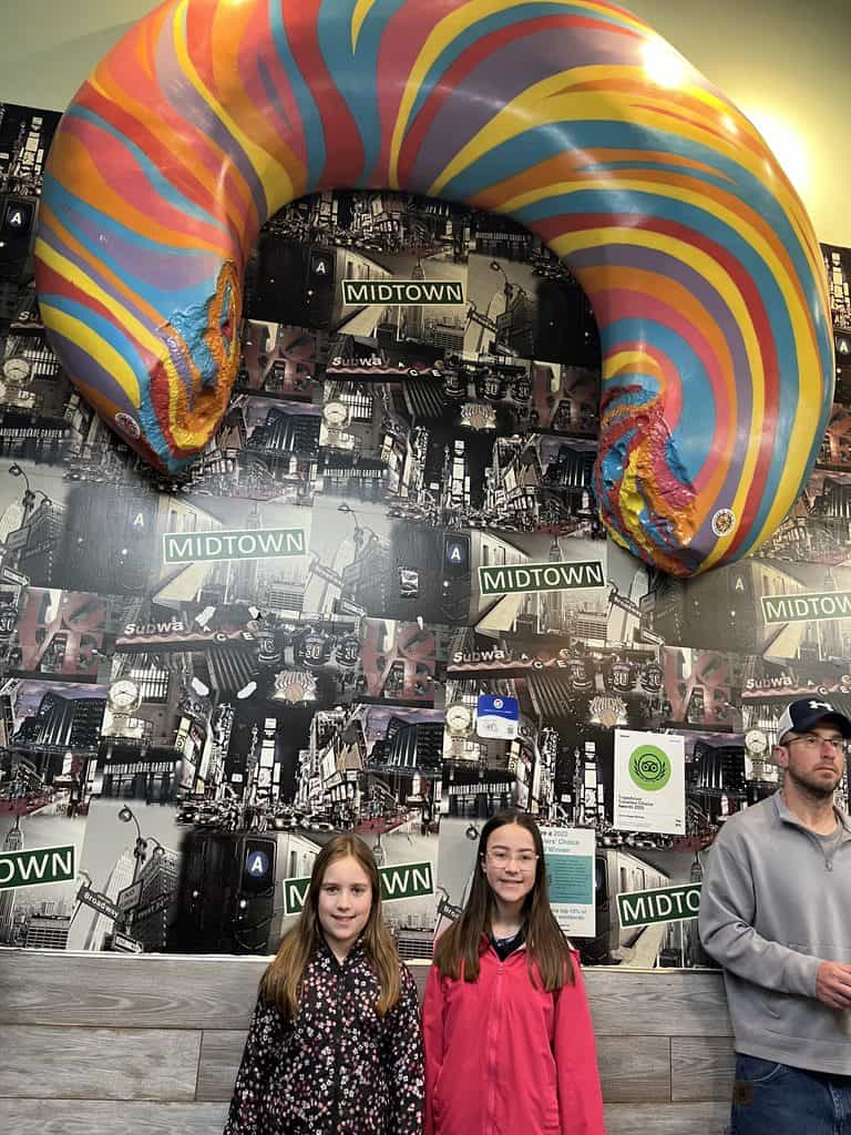 A family waits for rainbow bagels that kids love in NYC. Two girls smile below a large rainbow bagel on the wall at a NYC bagel shop.