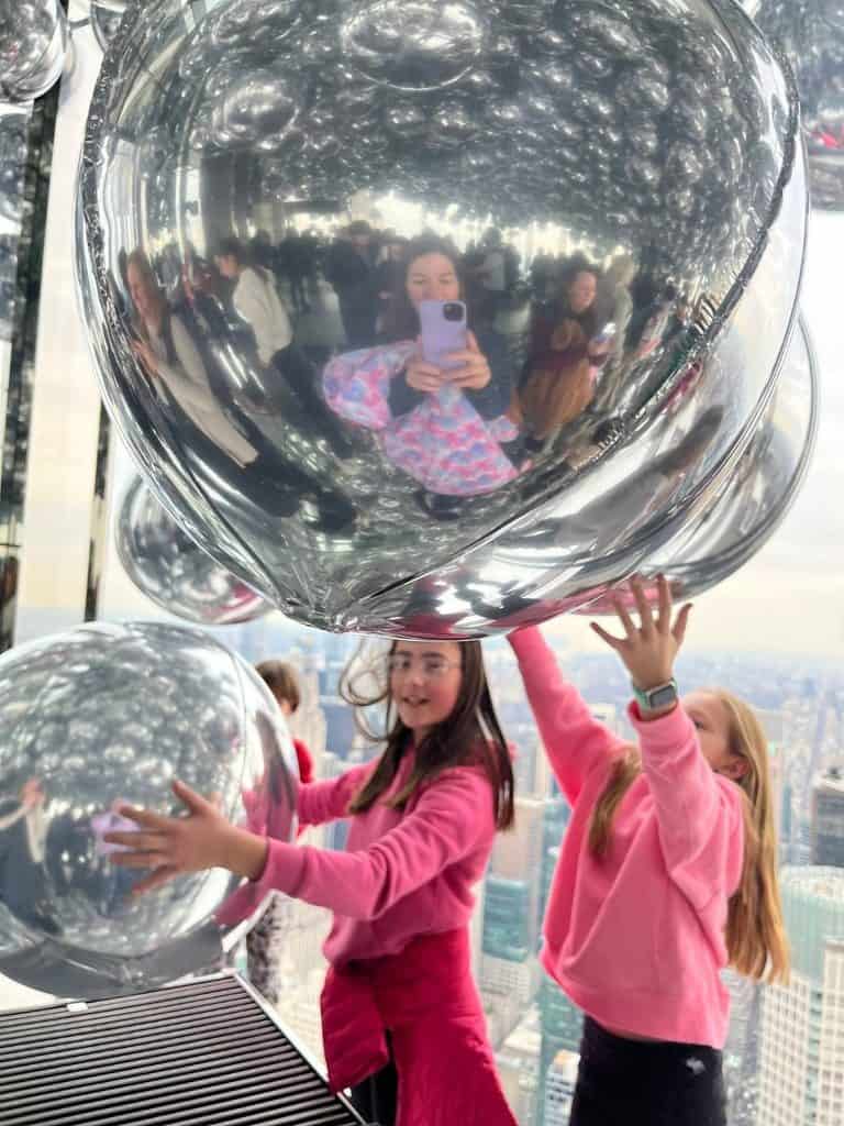 Two families visit NYC together and see Summit One. A mom takes a photo of a girl and her friend at Summit One's silver balloon room.