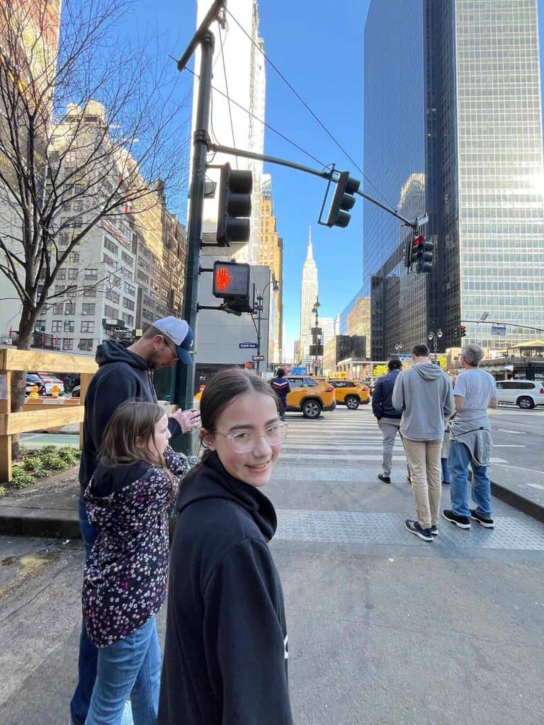 A family visits NYC for the first time. A girl smiles while walking on a street toward the Empire State Building while her family navigates NYC.