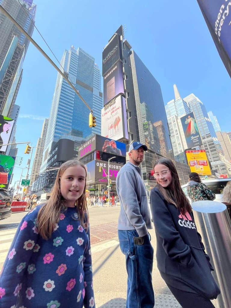 A family visits Times Square on their first NYC trip. A family smiles in NYC's Times Square with colorful billboards and tall buildings.